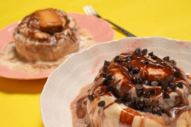 Zengin çikolata sosuyla kaplı taze tarçınlı çöreğin nefis bir yakın çekimi ve arka planda başka bir hamur işi ile mini siyah çikolata parçacıkları serpiştirilmiş.