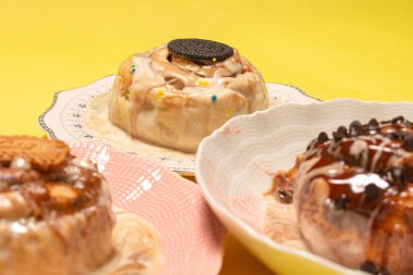 Farklı tatlı soslu üç gurme tarçınlı çöreğin karışımı. Kurabiyeye özel bir odak noktası var ve sarı arka planda pasta serpiştiriyor.