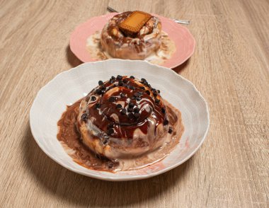 İki gurme tarçınlı çöreğin tabakta farklı malzemelerle sunulduğu ön çikolata parçalı hamur işine odaklanmış açık ahşap bir masa.