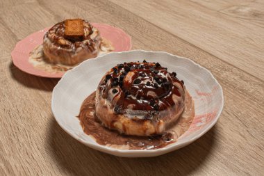 İki farklı lezzetli tarçınlı rulo, biri bisküvili, diğeri de renkli tabakta çikolata parçalı, ahşap bir masada bir şeyler atıştırmaya hazır.