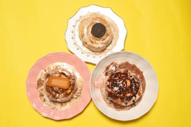 Renkli tabaklarda üç gurme tarçınlı çöreğin, parlak sarı bir arka planda farklı tatlı malzemelerle üst üste sunumunun