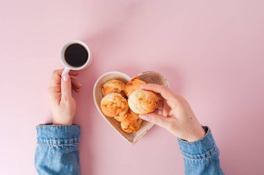 Kahve fincanı ve kalp şeklinde bir kaseyle yumuşak pembe arka planda Brezilya peynirli ekmeği tutan bir kız.