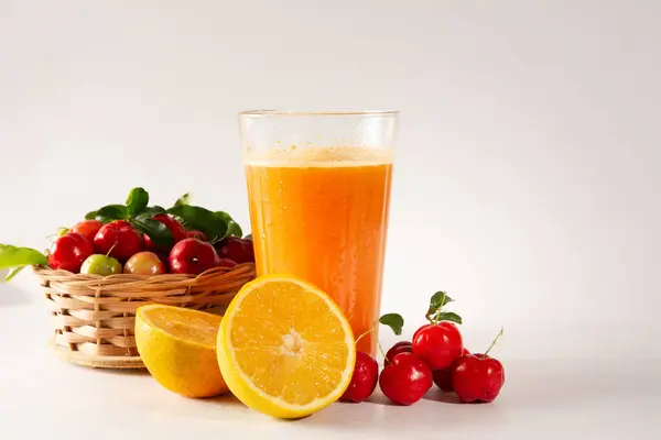 stock image Fresh Organic Acerola and Orange Juice in a glass cup with half orange and acerola berries inside a wood basket in a white clean studio photo in front view