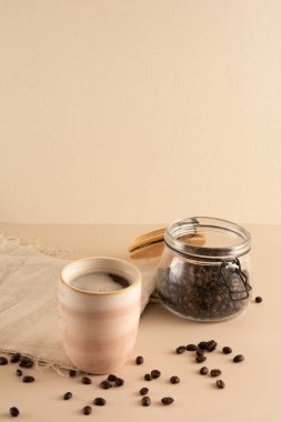 Kahve fincanı, kahve çekirdekleri ve masa örtüsüyle bej arka planda.