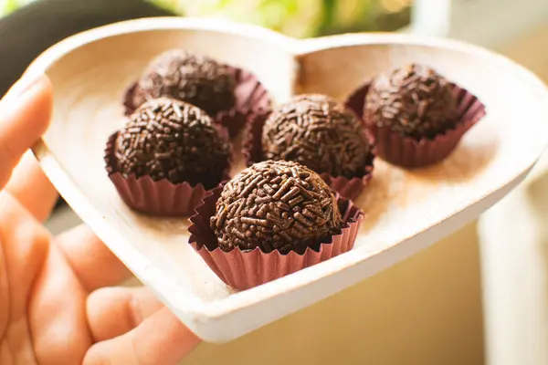 stock image Brazilian chocolate truffles (brigadeiros), a chocolate ball candy , on a heart-shaped plate, held by a woman's hand, outside. Party birthday candy, close up, sun light