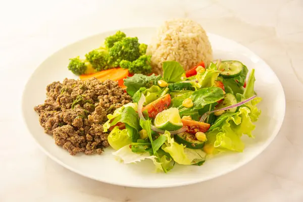 stock image This image showcases a nutritious meal consisting of seasoned ground beef, steamed broccoli, sliced carrots, brown rice, and a fresh mixed salad with greens, cherry tomatoes, red onions, and lime wedges. Perfect for a balanced diet, this plate offers