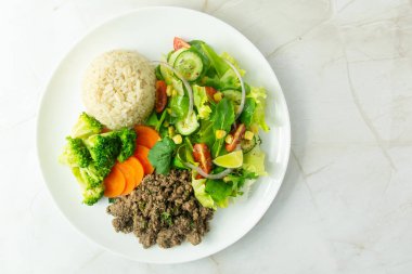 İçinde kıyma, esmer pirinç, buharda brokoli ve havuç ve domatesli taze salata, salatalık ve beyaz tabakta mısır bulunan sağlıklı bir yemek..