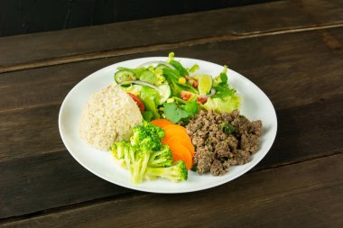 İçinde kıyma, esmer pirinç, buharda brokoli ve havuç ve domatesli taze salata, salatalık ve beyaz tabakta mısır bulunan sağlıklı bir yemek..