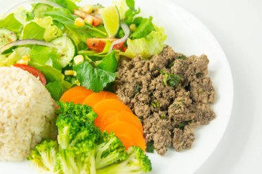 İçinde kıyma, esmer pirinç, buharda brokoli ve havuç ve domatesli taze salata, salatalık ve beyaz tabakta mısır bulunan sağlıklı bir yemek..