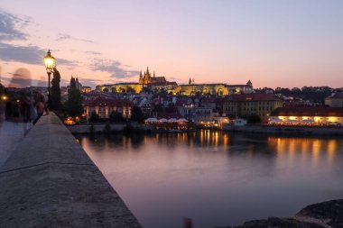 Charles Köprüsü 'nden Prag Kalesi' nin alacakaranlık manzarası. Gün batımı renkleri sakin Vltava Nehri 'ni yansıtıyor. Siluetli kuleler parlayan gökyüzüne karşı yükselir, dingin ve romantik bir atmosfer yaratır..