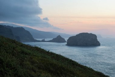 Matxitxako Deniz Feneri 'nden San Juan de Gaztelugatxe Kilisesi' nin sakin bir alacakaranlık manzarası. Önünde engebeli uçurumlar ve yemyeşil alanlar var..
