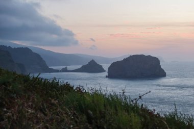 Matxitxako Deniz Feneri 'nden San Juan de Gaztelugatxe Kilisesi' nin sakin bir alacakaranlık manzarası. Önünde engebeli uçurumlar ve yemyeşil alanlar var..