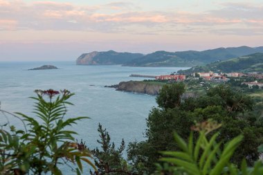 Bermeo 'nun Bask Ülkesi, İspanya' daki manzarası. Ağaçlar tarafından çerçevelenen günbatımı gökyüzünde pembe renkler yaratırken, yeşil tepeler ve uçurumlar okyanusun masmavi sularına sınır çiziyor..