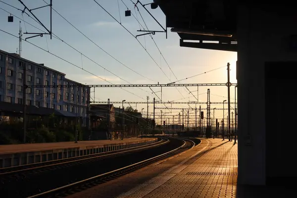 Prag Liben tren istasyonunda gün batımında, elektrik hatlarından oluşan bir ağ altında raylar kıvrılıyor. Ilık ışık platformun üzerinde uzun gölgeler oluşturarak istasyonun endüstriyel mimarisini vurguluyor..