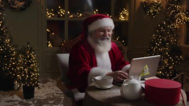 Meşgul Noel Baba dizüstü bilgisayarında çalışıyor, bazı notlar ve planlar yapıyor, mutlu ve gülümsüyor, tatil öncesi dönemin tadını çıkarıyor, Ağır çekim.