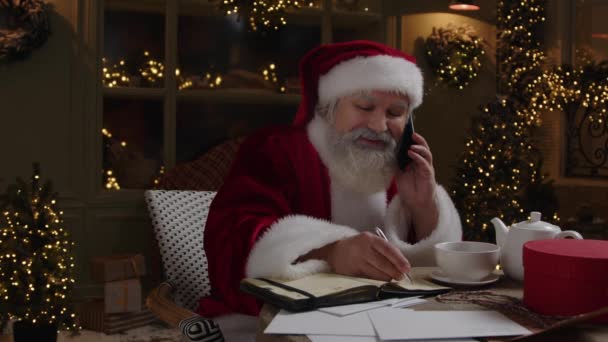 サンタクロースは夜遅く 携帯電話を話し いくつかの休日の手配をして ノートブックにメモを作る スローモーション — ストック動画