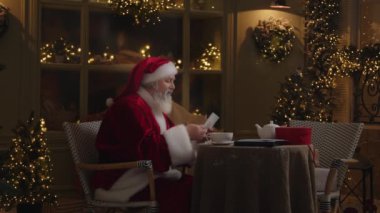 Noel Baba evdeki rahat arka bahçede çalışıyor, ışıklarla ve Noel ağaçlarıyla süslenmiş, zevkle çocukların mektuplarını okuyor ve çay içiyor, sihirli kış sezonu, yavaş çekim..