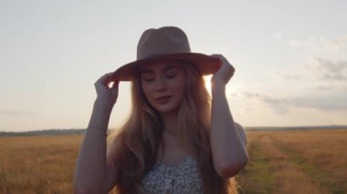 Şapkalı genç bir kız, güzel bir yaz günbatımının arka planında kameraya bakar. Ağır çekim. Yakın plan.