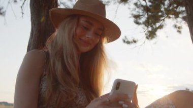 Güzel bir yaz günbatımının arka planında bir ağacın altında oturan şapkalı genç bir kız akıllı telefon kullanır. Yavaş hareket. Yakın çekim.