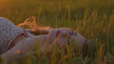 Genç ve güzel bir kız yaz günbatımının arka planında çimlerin üzerinde uzanır ve kameraya bakar. Ağır çekim. Yakın plan.
