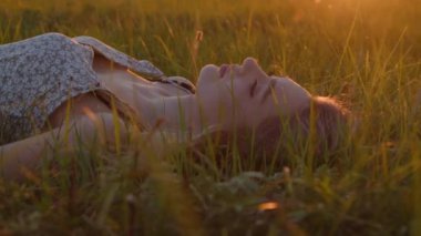 Genç ve güzel bir kız yaz günbatımının arka planında çimlerin üzerinde yatıyor. Ağır çekim. Yakın plan.