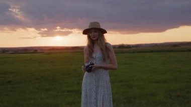 Şapkalı genç bir kız elinde antika bir fotoğraf makinesi tutuyor ve güzel bir yaz günbatımının arka planında bir fotoğraf çekiyor.