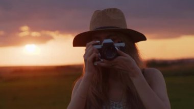Şapkalı genç bir kız elinde antika bir fotoğraf makinesi tutuyor ve güzel bir yaz günbatımının arka planında bir fotoğraf çekiyor.