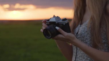 Şapkalı genç bir kız elinde antika bir fotoğraf makinesi tutuyor ve güzel bir yaz günbatımının arka planında bir fotoğraf çekiyor.