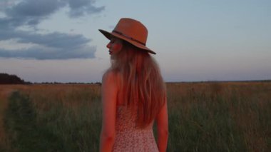 Şapkalı genç bir kız güzel bir yaz günbatımının arka planında koşup kameraya bakar. Ağır çekim. Yakın plan.