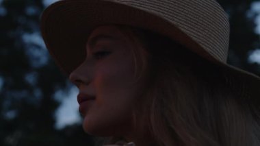 Şapkalı genç bir kız, güzel bir yaz günbatımının arka planında kameraya poz veriyor ve kameraya bakıyor. Ağır çekim. Yakın plan.