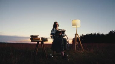 Genç bir kadın klasik bir lambanın yanında oturuyor ve alacakaranlıkta bir kitap okuyor, güzel bir siluet, geniş çekim, yüksek kalite 4K görüntü.