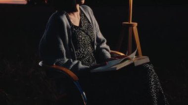 Jumper giymiş genç bir kadın dışarıda oturmuş kitap okuyor, hobisinin tadını çıkarıyor, yaz akşamı, alacakaranlık, klasik tarz, yüksek kalite 4K görüntü.