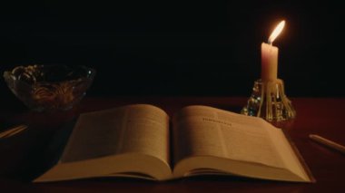 Masanın üzerinde açık bir kitap, mum ışığında İncil, yakınlarda mum yanıyor, ruhani uygulama, yüksek kaliteli 4K görüntü.