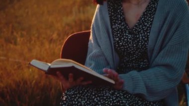 Gri kazaklı ve gözlüklü genç bir kadın sahada oturuyor ve bir roman okuyor, tek başına rahatlıyor, güzel altın gün batımı, yakın çekim, yüksek kalite 4K görüntü