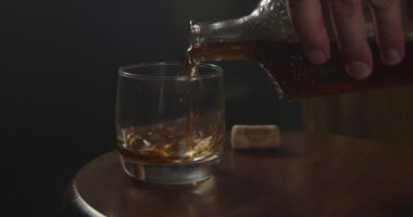 Yakından bakınca, bir elin maharetli bir şekilde koyu renkli bir sıvıyı bardağa doldurduğunu görüyoruz. Yanında da bir mantar duruyor.