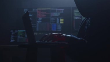 Yetenekli bir hacker 'ın karanlık silueti, seyrek aydınlatılmış bir ortamda yoğun bir şekilde dizüstü bilgisayarla çalışırken görülüyor.