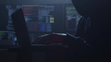 Yetenekli bir hacker 'ın karanlık silueti etraftaki birden fazla parlayan ekranın arasında aktif olarak dizüstü bilgisayarda yazıyor.