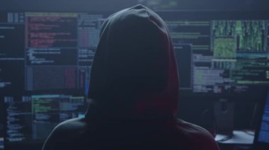 Belirsiz ışıklandırılmış bir ortamda kod ve veri sinyallerini gösteren birden fazla ekrana bakan kapüşonlu bir figür.