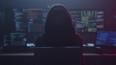 Kapüşonlu bir hacker 'ın gölgeli silueti çeşitli kodlar içeren parlak ekranlarla çevrilidir.