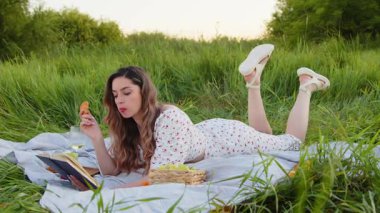 Genç bir kadın mutlu bir şekilde dışarıda piknik yapıyor, kitabını okuyor ve yemyeşil bir tarlada atıştırıyor.