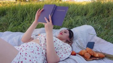 Bir kadın çimenlerin üzerinde rahat bir battaniyenin üzerinde büyüleyici bir kitap okuyor, güneşli bir günün tadını çıkarıyor hafif bir esintiyle