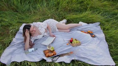 Sakin bir çimenlikte, bir kadın taze meyvelerin tadını çıkarırken büyüleyici bir kitap okumaktan zevk alır.