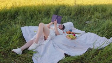 Bir kadın, doğada bir piknik battaniyesinde otururken kitap okumanın zevkini çıkarıyor.