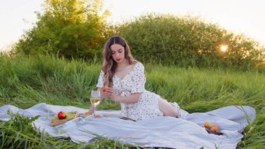 Huzurlu bir piknik sahnesinde bir kadın çeşitli meyvelerden ve açık havada güzel bir bardak şaraptan keyif alır.