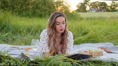 Açık havada mutlu bir şekilde kitap okuyan, şarabın tadını çıkaran ve lezzetli atıştırmalıkların tadını çıkaran bir kadınla huzurlu bir piknik sahnesi.