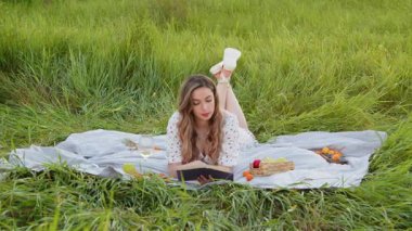 Huzurlu ve rahat bir piknik sahnesi. Resimli yeşil bir tarlada atıştırırken zevkle kitap okuyan bir kadın.