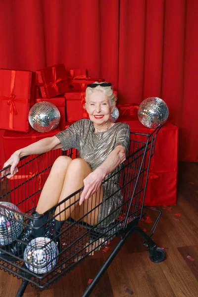 Senior stylish gray haired woman in sunglasses and silver dress sitting in the supermarket cart at the party, drinking wine on red curtains background. party, disco, celebration, senior age concept