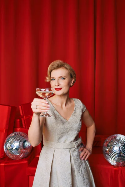 mature stylish elegant woman in cocktail dress with glass of sparkling wine with presents on red background. Party, fashion, celebration, anti age concept