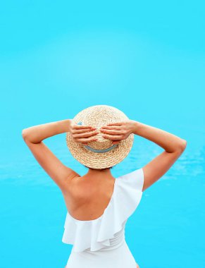 Beyaz mayo giymiş beyaz bir kadın yaz günü hasır şapkayla havuz kenarında (ya da deniz ya da okyanus) oturuyor. Yaz, rahatlama, sağlık, dinlenme konsepti