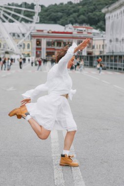 Güzel, mutlu bir kadın. Beyaz elbiseli, arka planda dönme dolap olan şehir meydanında zıplıyor.. 